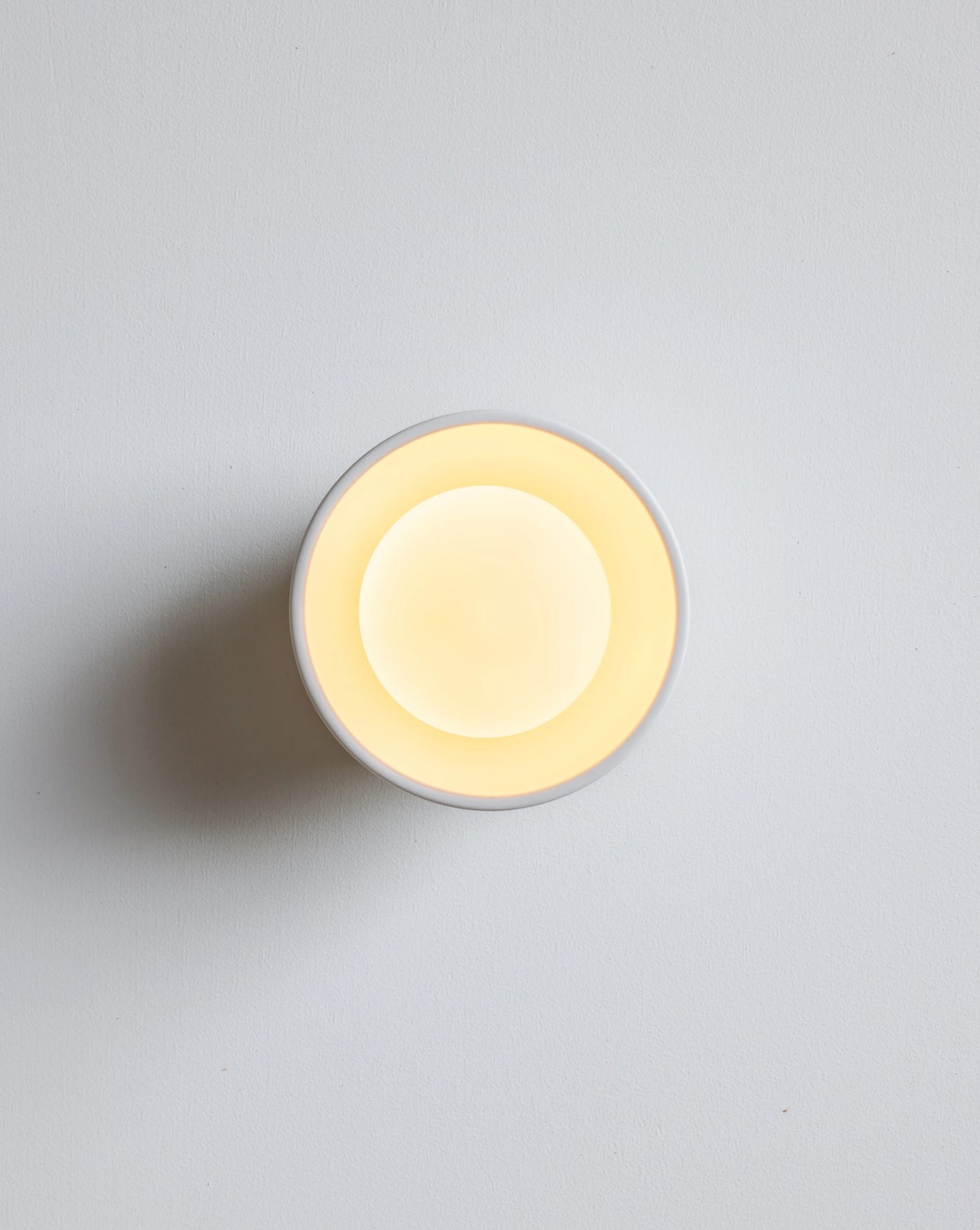 A minimalist round wall sconce with a white outer rim and a glowing warm yellow inner light, photographed against a clean light gray wall.