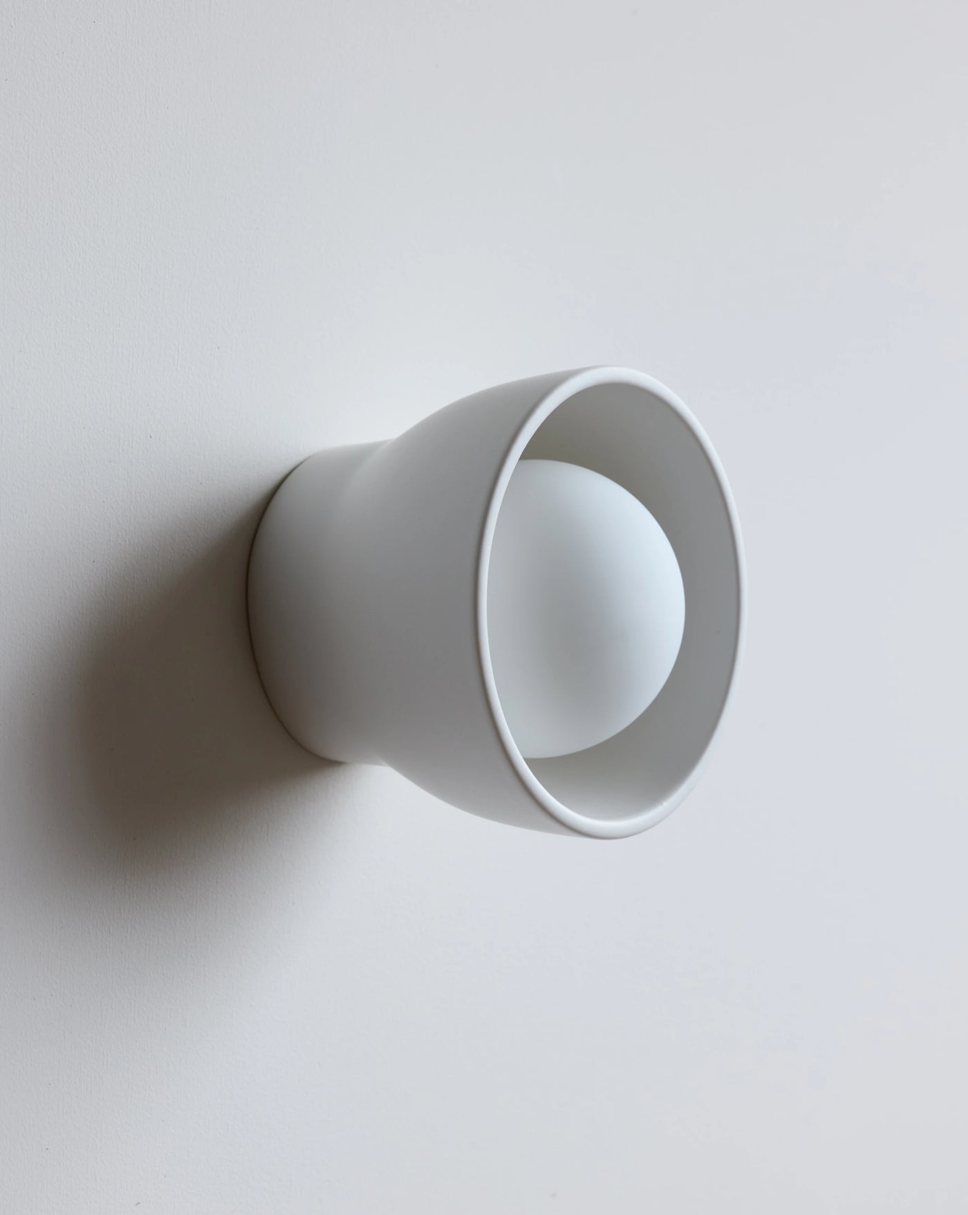 A minimalist white wall-mounted light fixture with a wide flared rim and a spherical bulb inside, shown against a plain light gray wall.