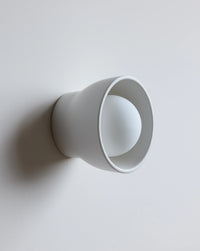 A minimalist white wall-mounted light fixture with a wide flared rim and a spherical bulb inside, shown against a plain light gray wall.