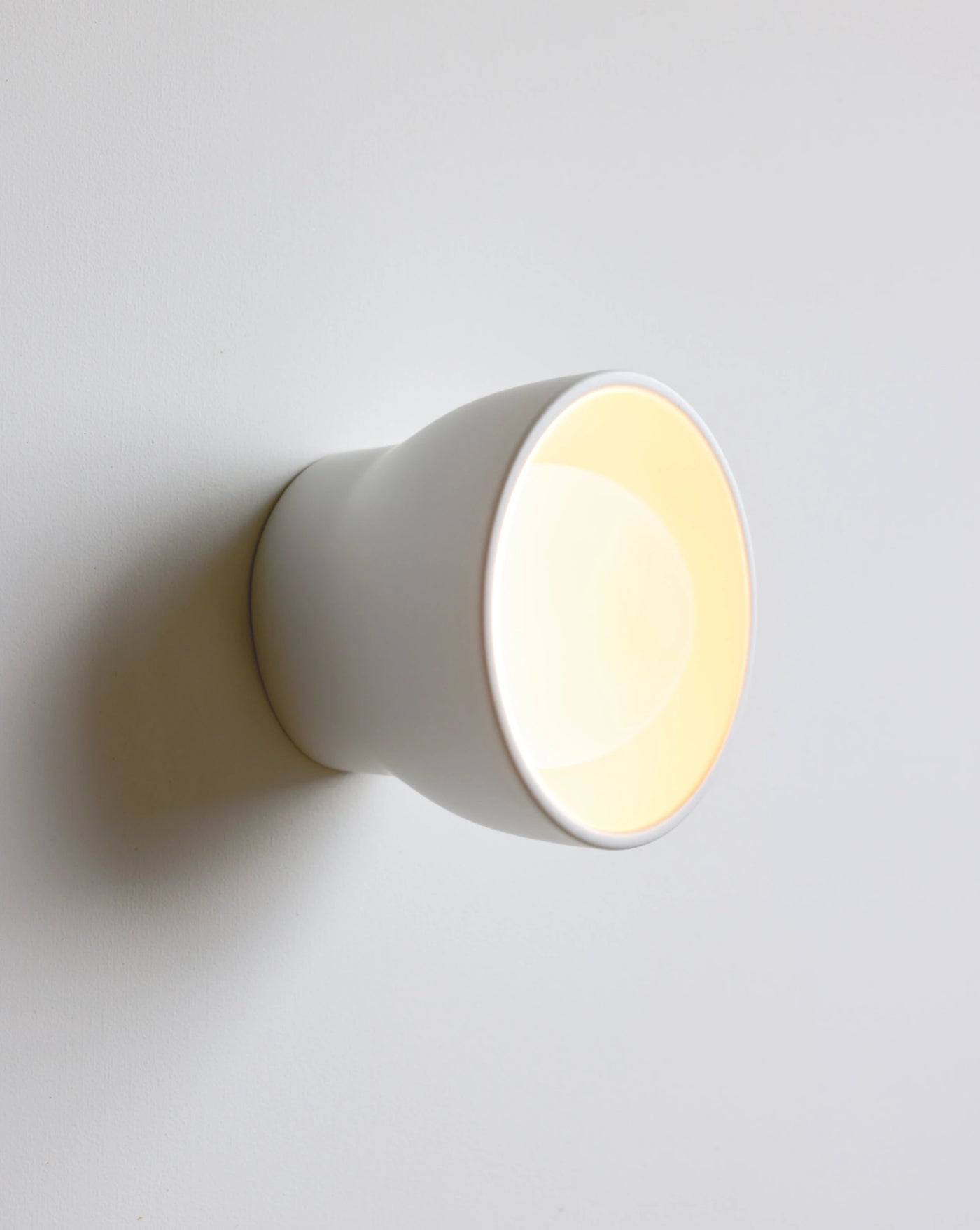 A minimalist white ceramic wall sconce light fixture, shown illuminated with a warm yellow glow against a clean, light gray wall.