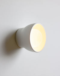 A minimalist white ceramic wall sconce light fixture, shown illuminated with a warm yellow glow against a clean, light gray wall.