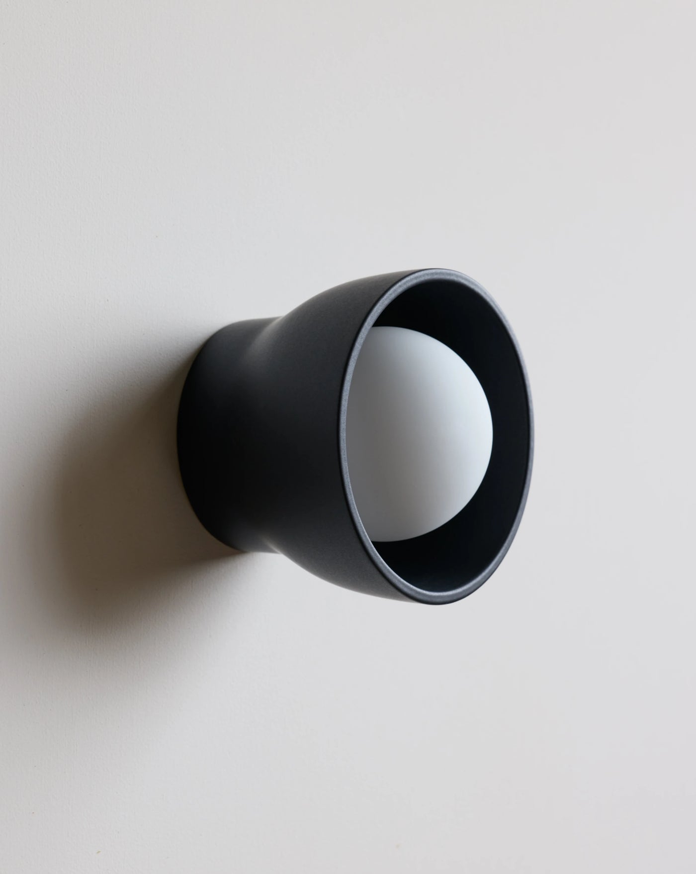 A modern matte black wall sconce with a wide flared shade and a white spherical bulb inside, mounted on a plain light gray wall.