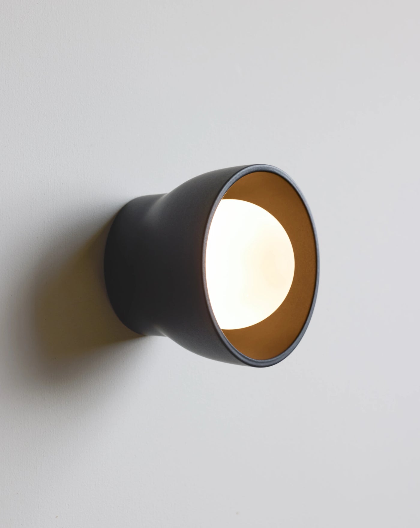 A modern wall-mounted sconce with a matte black exterior and a warm light bulb inside, casting a soft glow on a light gray wall.