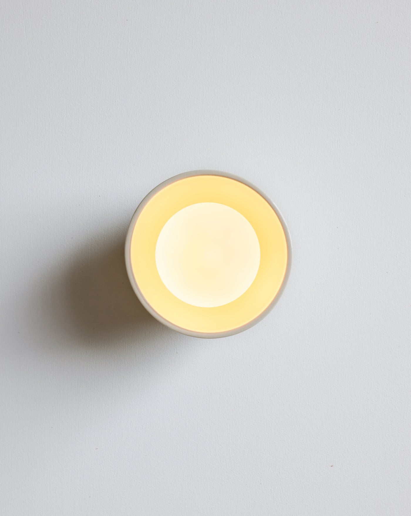 A minimalist round wall sconce light fixture with a warm yellow glow against a light gray wall.