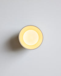 A minimalist round wall sconce light fixture with a warm yellow glow against a light gray wall.