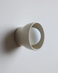 A minimalist wall-mounted light fixture with a matte off-white cone-shaped shade enclosing a spherical frosted bulb, shown against a plain light gray wall.