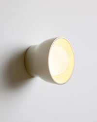 A modern wall-mounted cone-shaped light fixture in a warm cream finish, shown illuminated against a plain light gray wall.