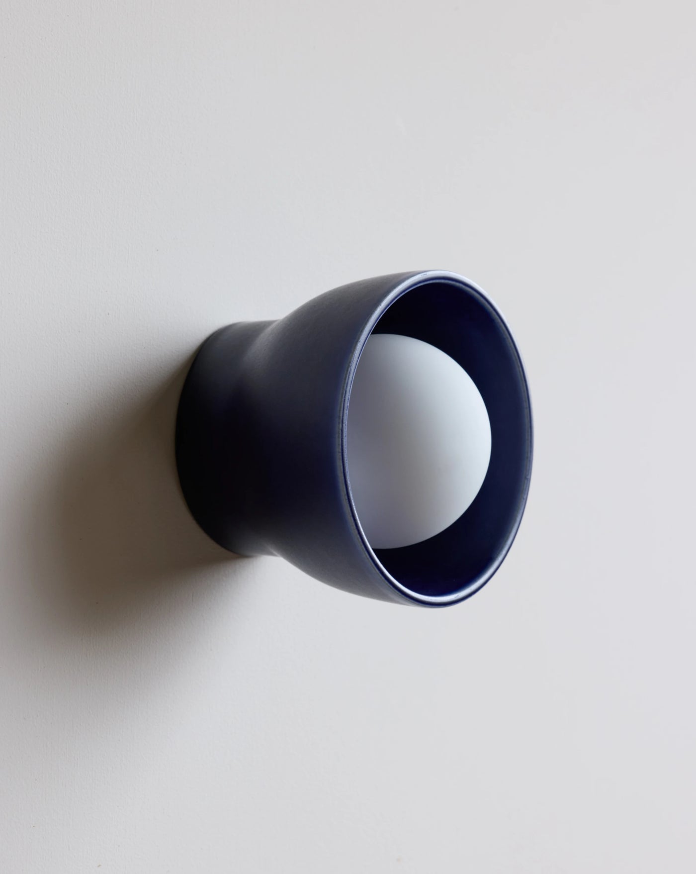 A modern wall-mounted light fixture with a matte navy blue cone-shaped outer shade and a white spherical bulb inside, shown against a plain light gray wall.