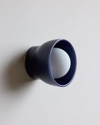 A modern wall-mounted light fixture with a matte navy blue cone-shaped outer shade and a white spherical bulb inside, shown against a plain light gray wall.