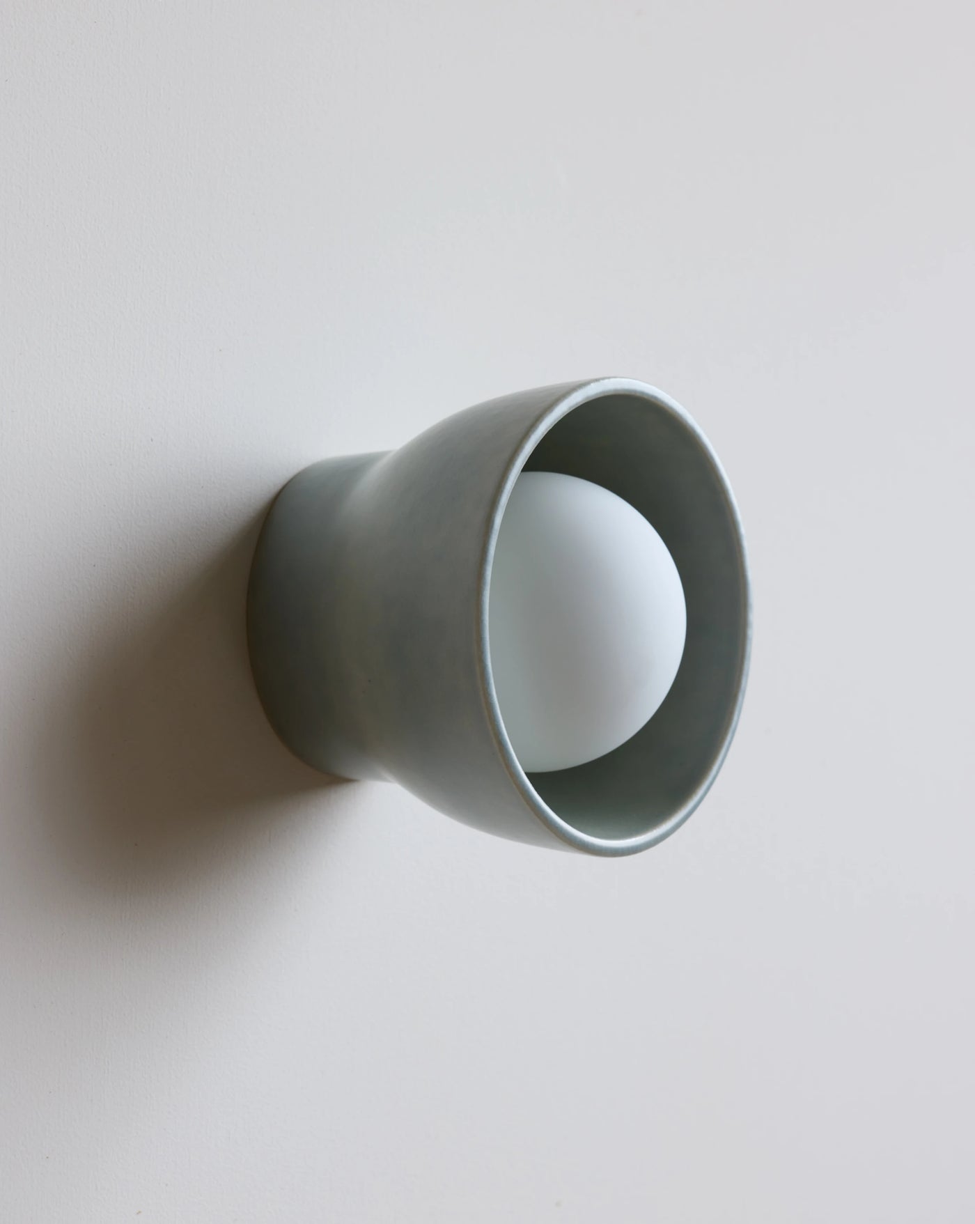A minimalist wall-mounted light fixture with a matte sage ceramic outer shade and a white frosted glass bulb inside, shown against a plain light gray wall.