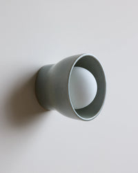 A minimalist wall-mounted light fixture with a matte sage ceramic outer shade and a white frosted glass bulb inside, shown against a plain light gray wall.
