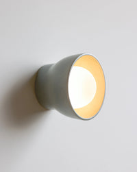 A modern wall-mounted light fixture with a matte sage exterior shown lit against a plain light gray wall.