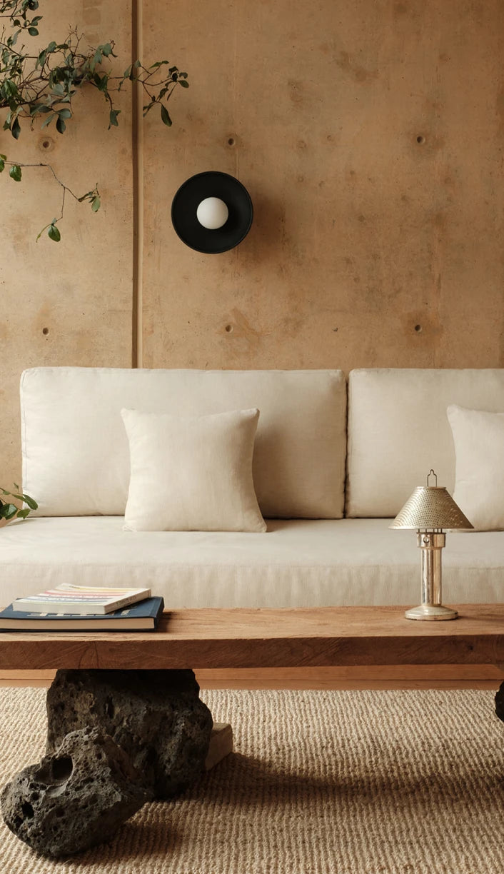 A minimalist living room scene featuring a cream-colored sofa with matching pillows, a rustic wooden coffee table with a dark rock base, and a textured beige wall with a black circular wall lamp.