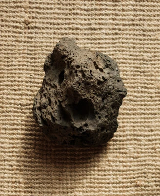 A rough, porous dark gray stone or volcanic rock piece resting on a textured beige woven fabric background.