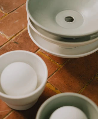 A stack of matte ceramic sconces in pale sage green and white and cream colorways, arranged on a rustic terracotta tile surface.