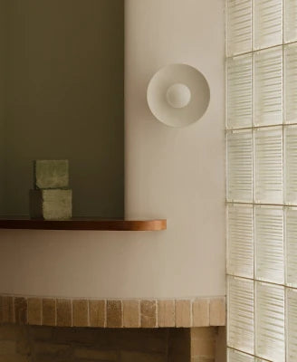 A minimalist white circular wall sconce mounted on a warm beige wall, positioned above a wooden shelf with stacked blocks, next to a glass block window.