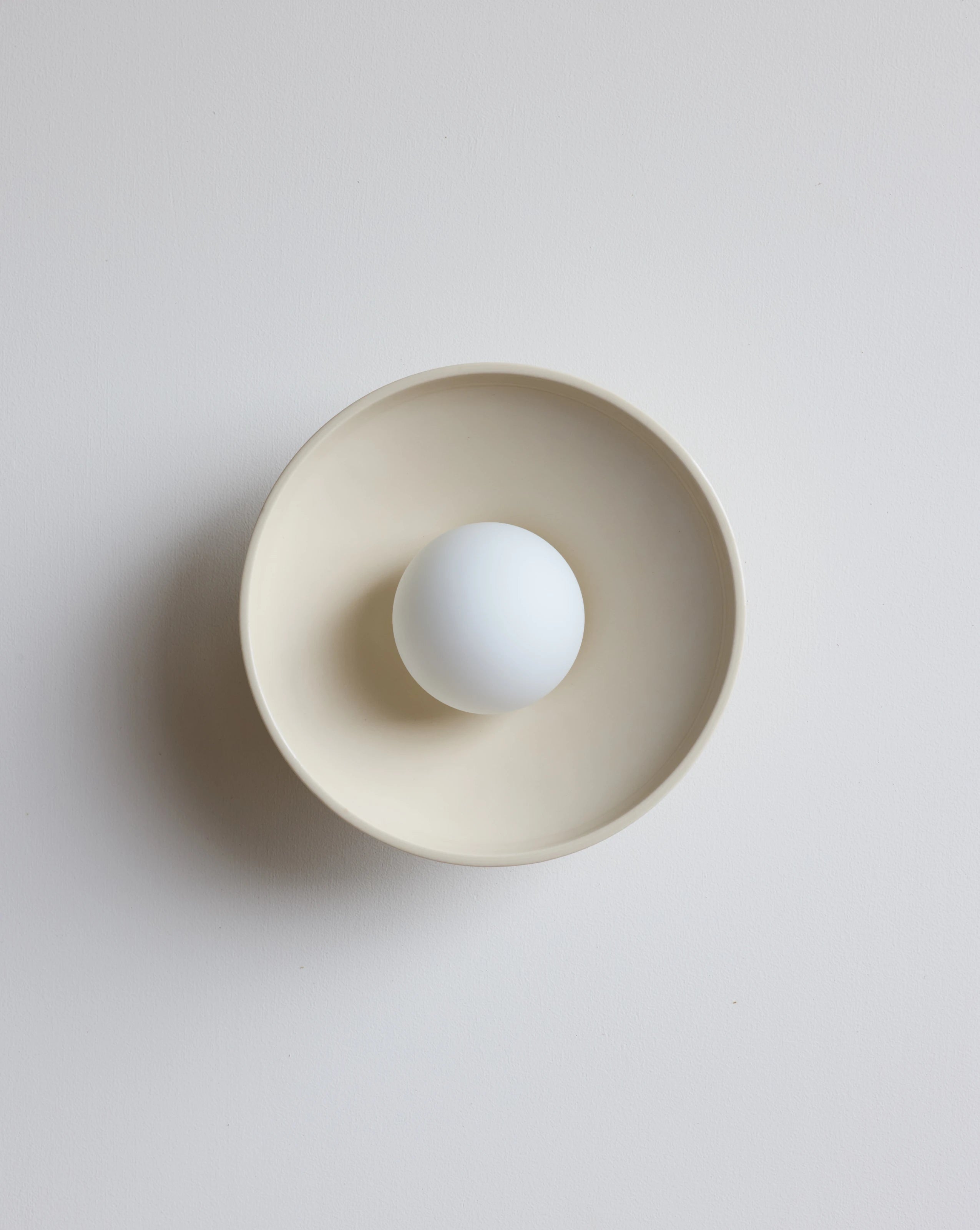 A minimalist ceramic sconce with a wide rim and a small white bulb centered inside, on a light gray background.