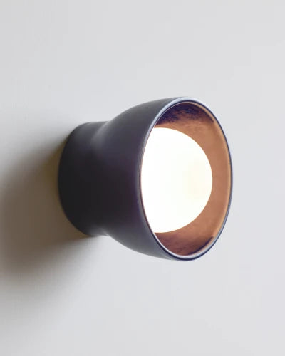 A modern wall-mounted sconce with a dark navy exterior casting a bright light against a neutral light gray wall.