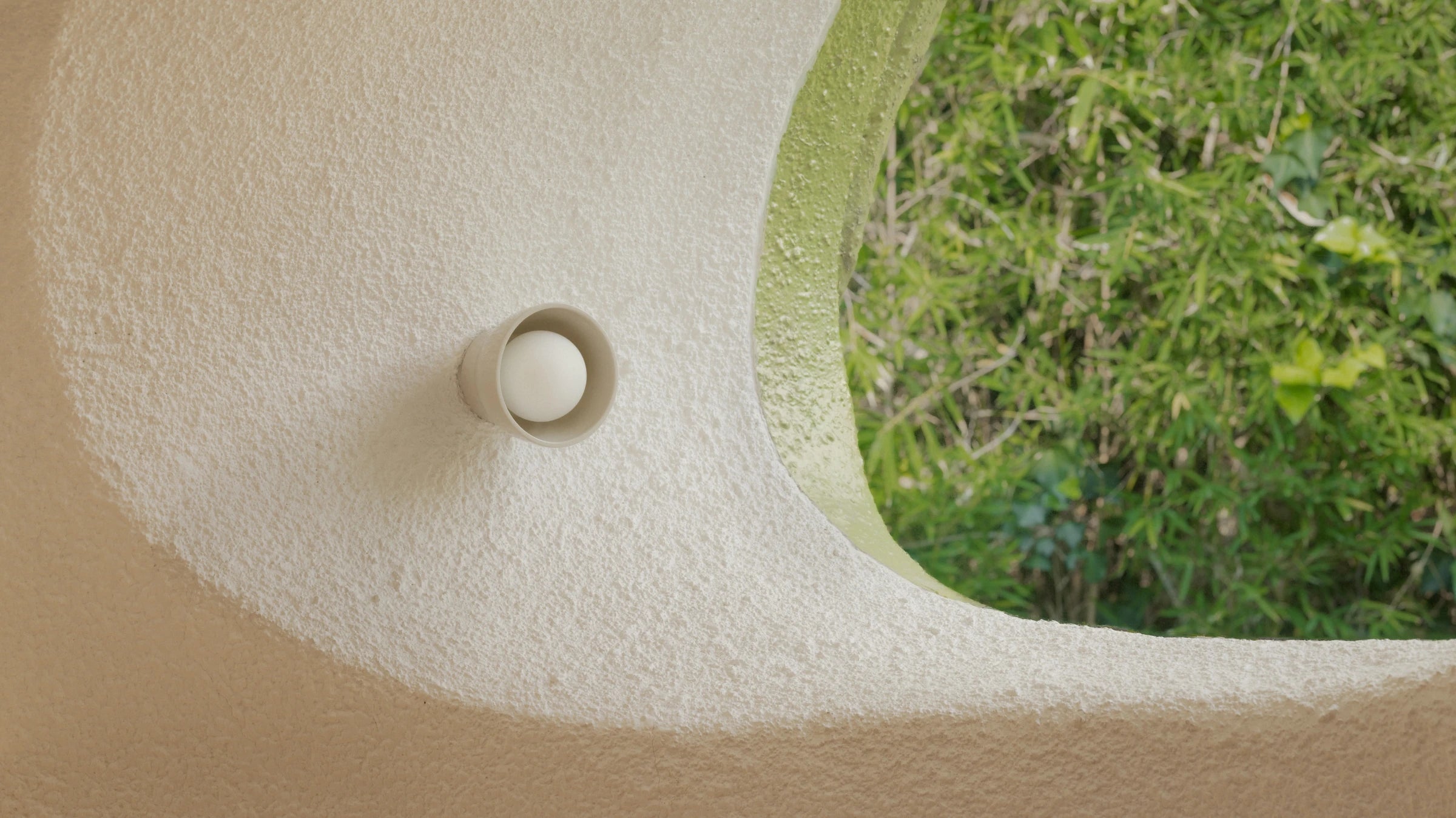 A close-up of a textured beige stucco wall with a mounted wall sconce, next to a curved opening showing green foliage outside.