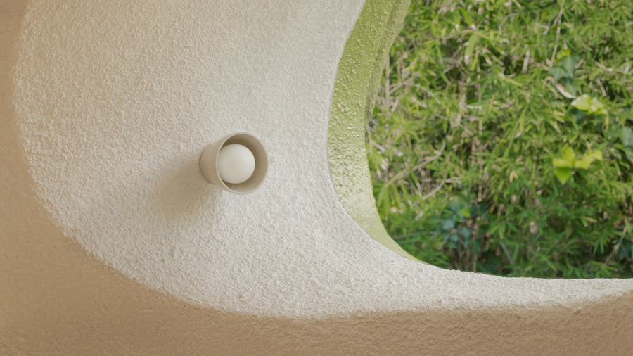 A close-up of a textured beige stucco wall with a mounted wall sconce, next to a curved opening showing green foliage outside.