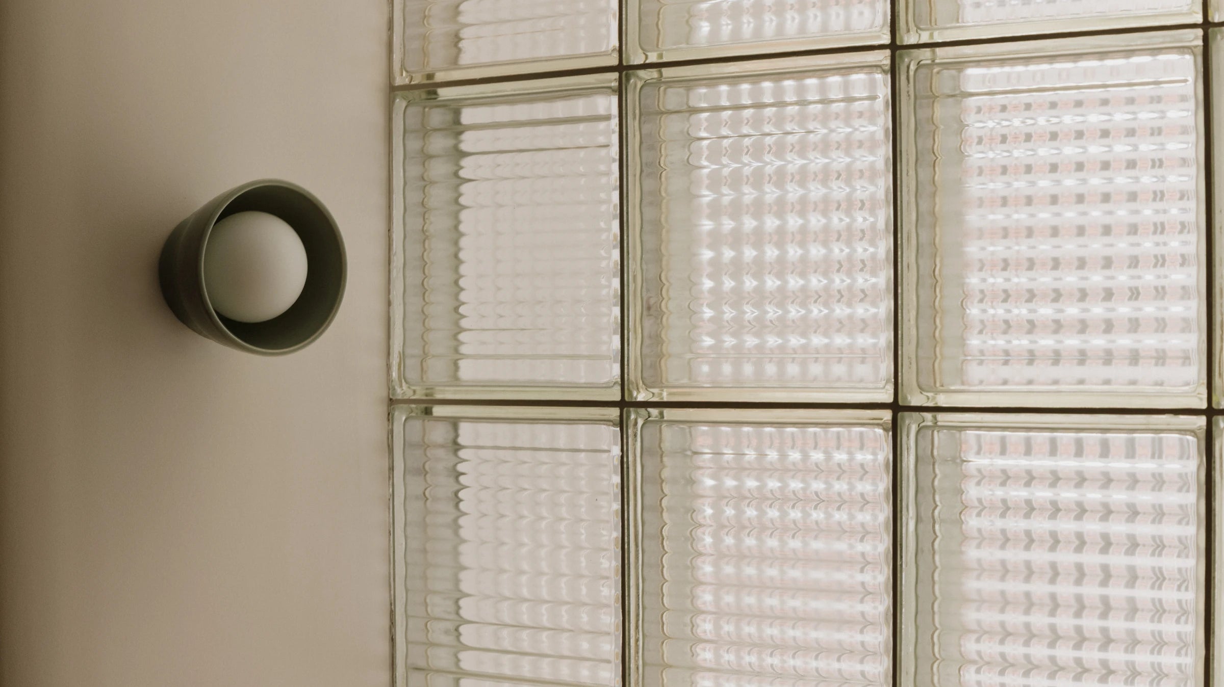 A modern matte cylindrical wall sconce mounted on a beige wall next to a glass block wall with textured frosted panels.