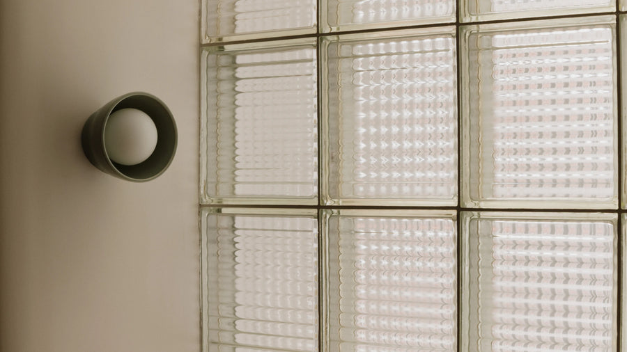 A modern matte cylindrical wall sconce mounted on a beige wall next to a glass block wall with textured frosted panels.