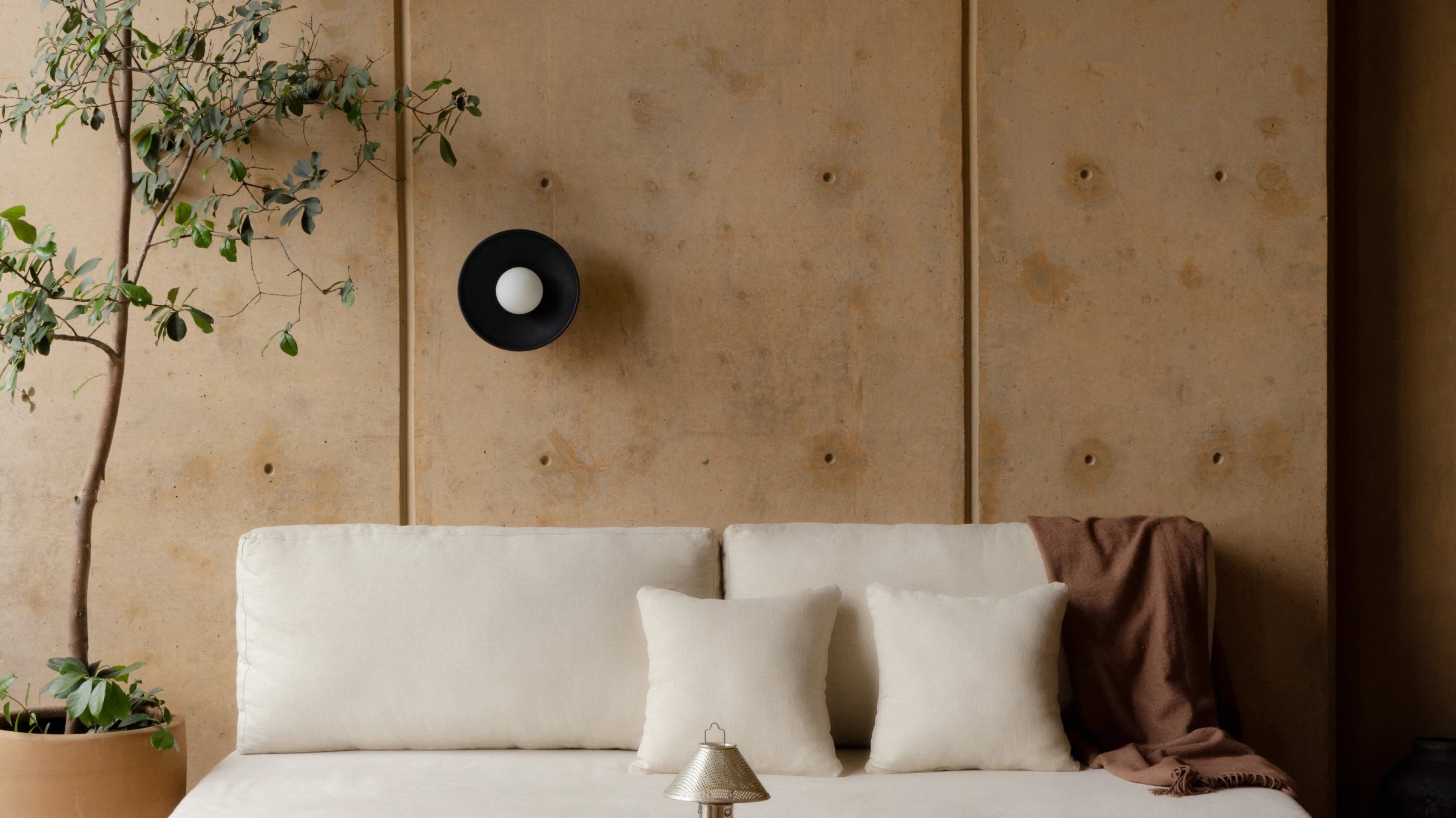 A minimalist bedroom scene featuring a low-profile white sofa with cream pillows and a brown throw blanket, set against a textured warm beige wall with a black circular wall sconce and a potted plant.