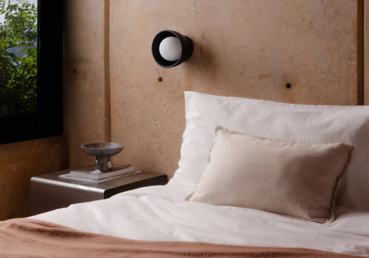 A minimalist bedroom scene featuring a neatly made bed with white linens and a beige accent pillow, set against a textured concrete wall with a black wall-mounted sconce.