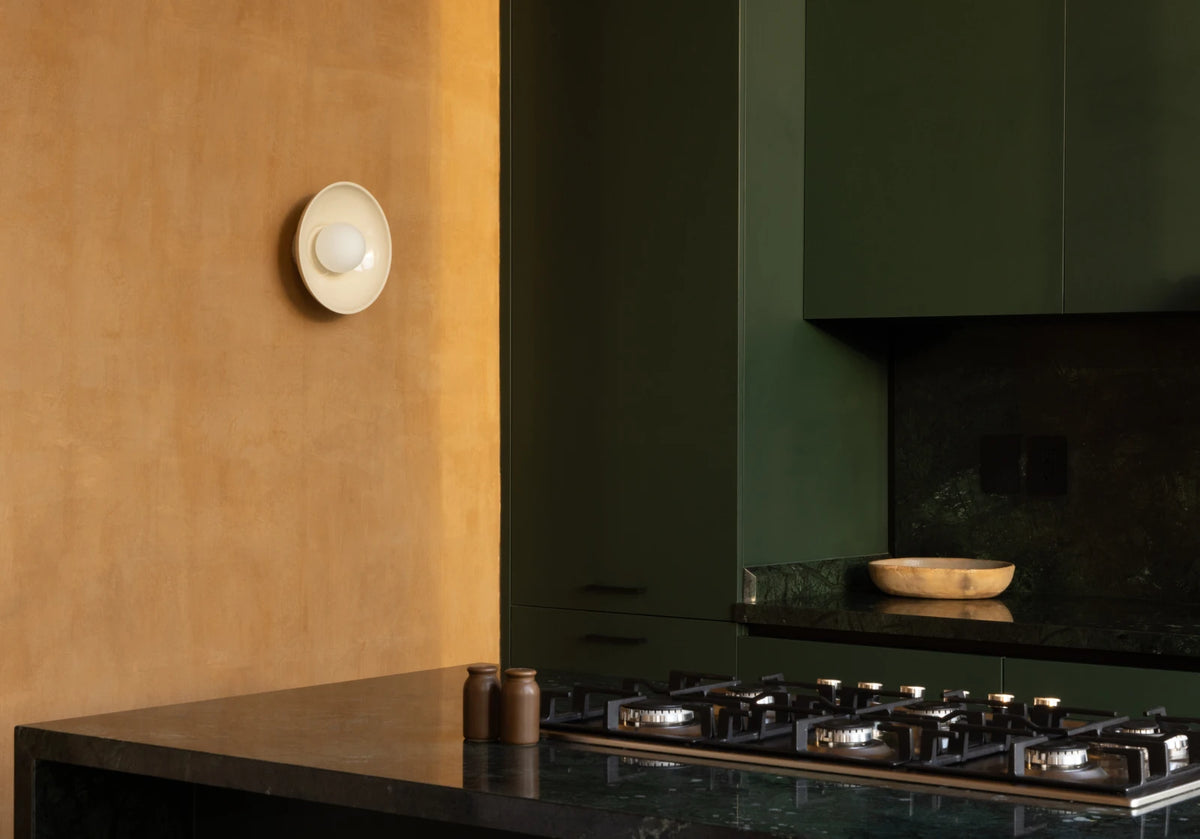A modern kitchen interior with dark green cabinetry, a black countertop and gas stovetop, and a warm plaster wall with a small white wall-mounted light fixture.