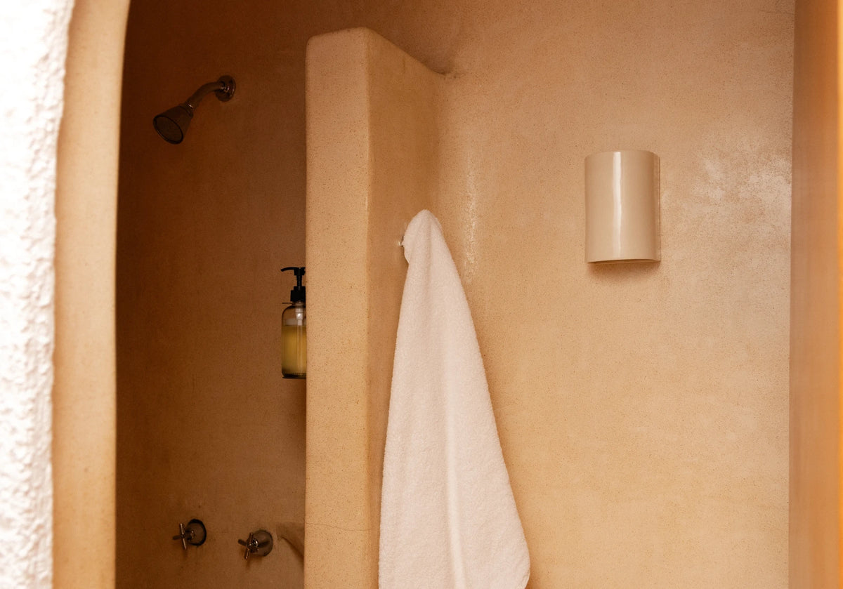 A warm-toned bathroom interior with a white towel hanging from a hook on a beige plaster wall, a shower head visible in the shower alcove, and a cylindrical wall light.