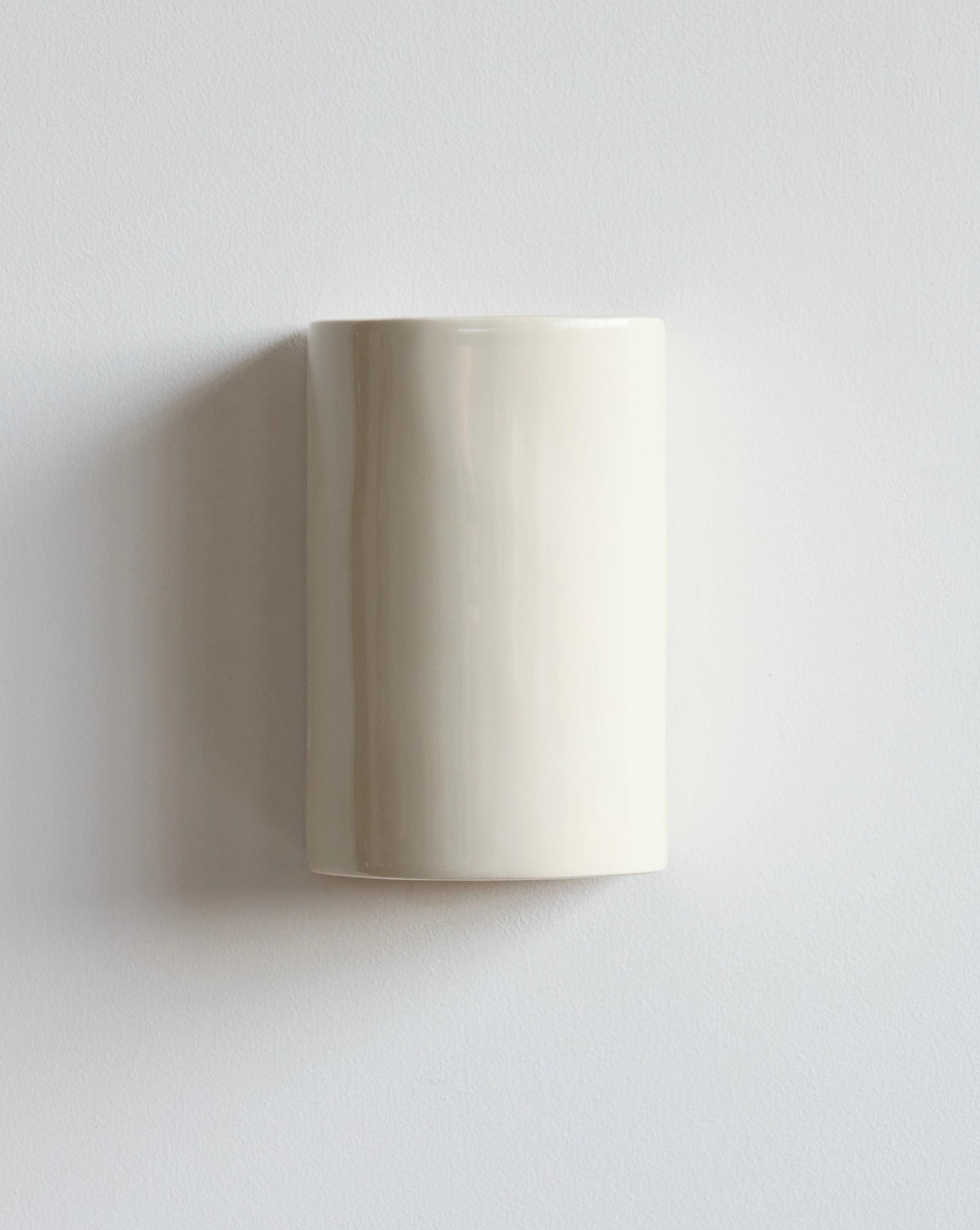 A minimalist, cylindrical wall sconce in a warm cream color, mounted on a plain light gray wall with a soft shadow to the left.