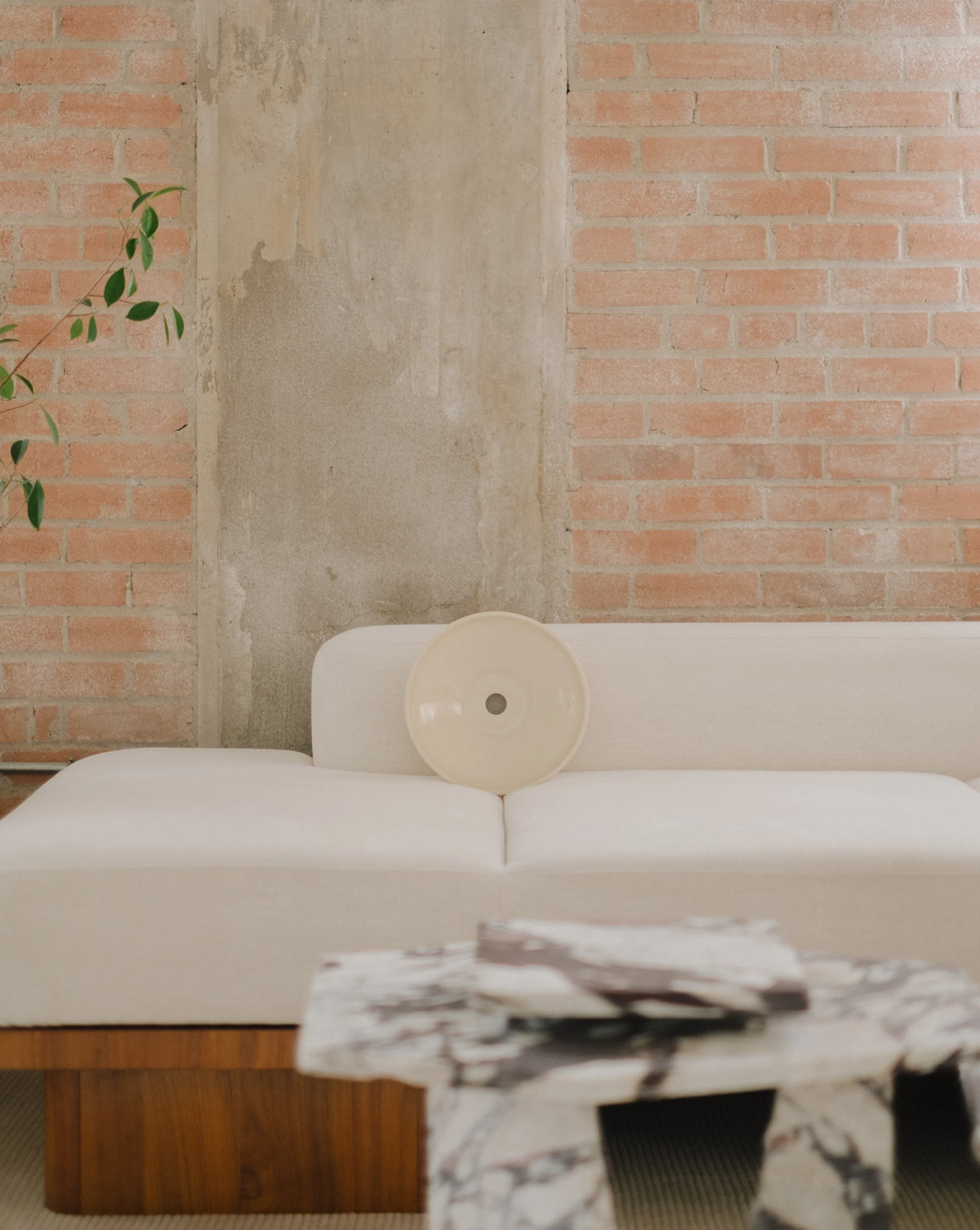 A cream-colored oval wall sconce resting on a white sofa in a modern living room setting with a brick wall background and a marble coffee table in the foreground.