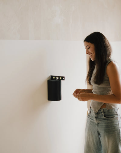 A woman in a gray sleeveless top and light blue jeans stands next to a black cylindrical wall-mounted light fixture on a white wall.