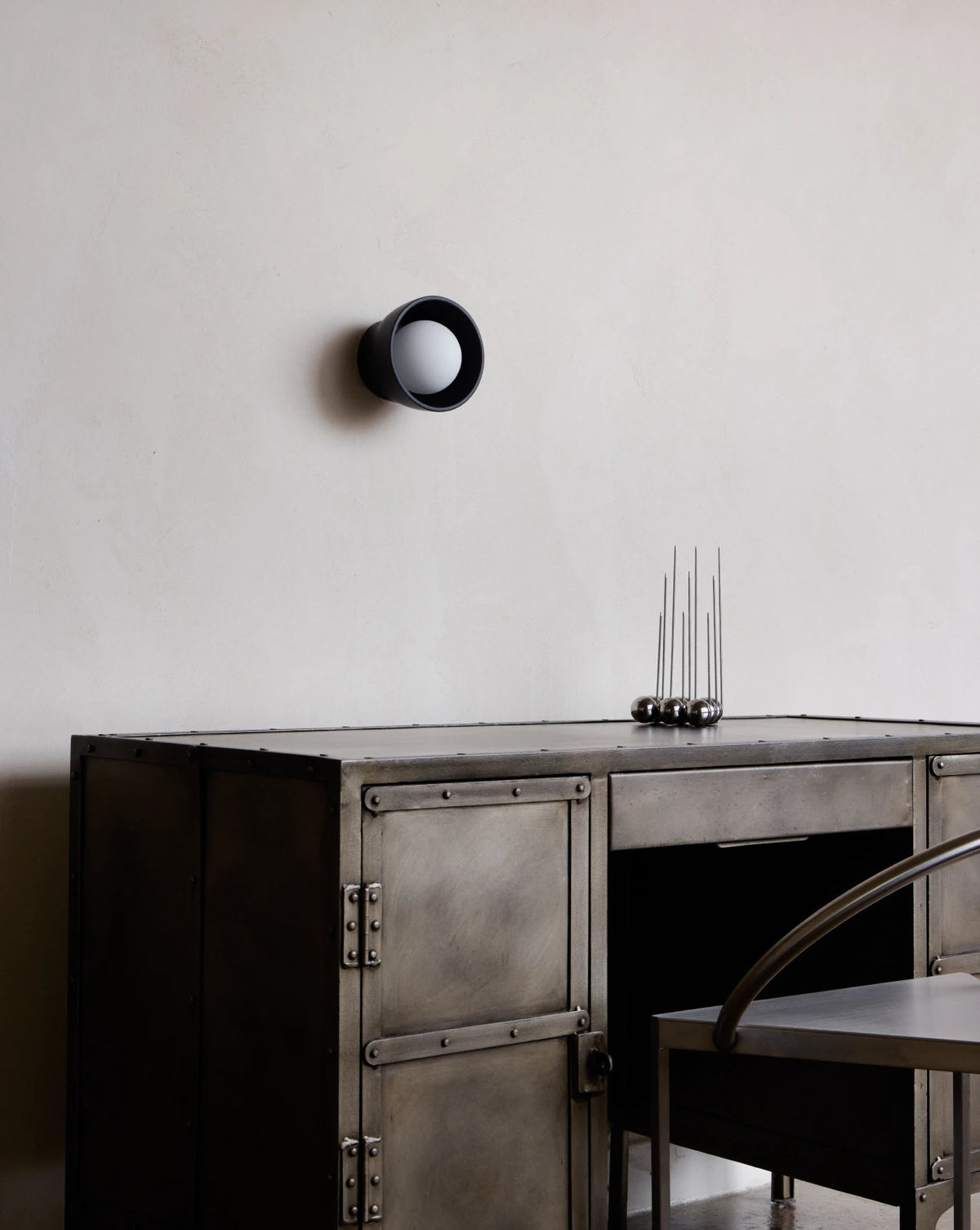 A dark metal industrial desk with rivets and a matching matte ceramic wall sconce mounted on a textured off-white plaster wall. A small sculpture sits on top of the desk.