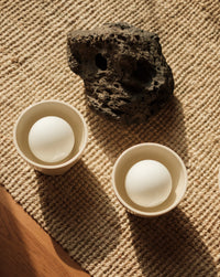 Two small beige ceramic wall sconces arranged on a woven beige rug beside a dark rough stone. A strip of wooden surface is visible in the lower left corner.