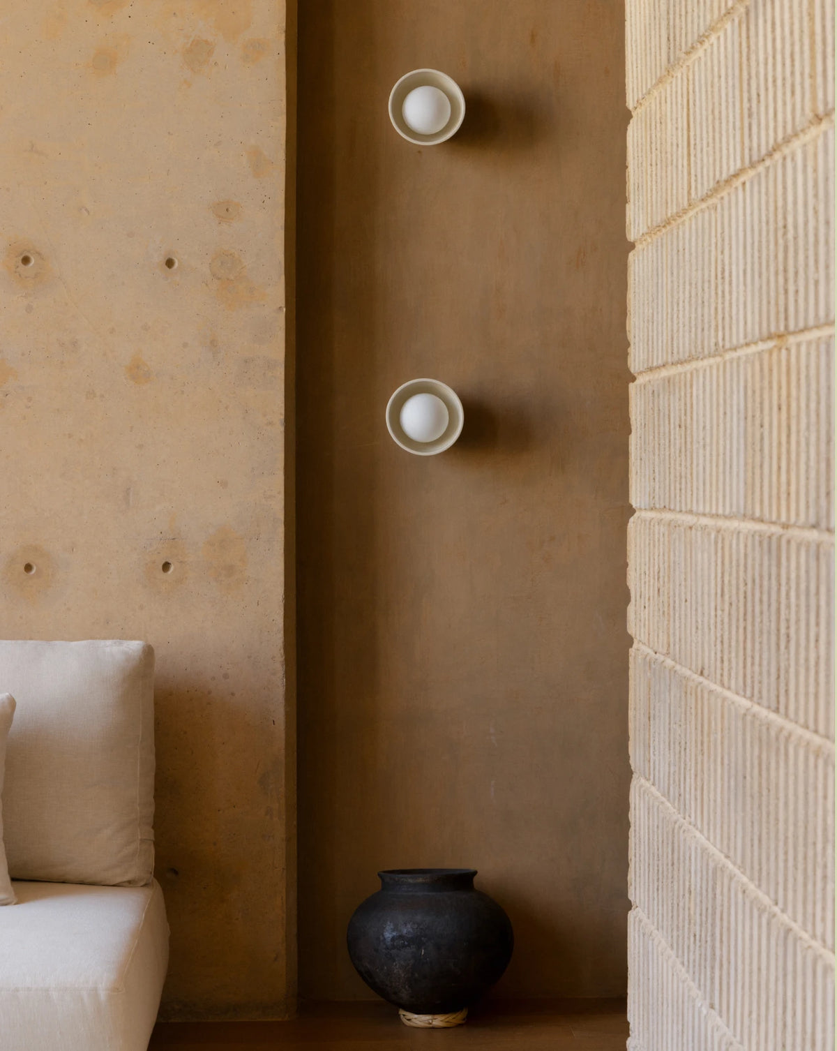 Two modern wall sconces with white circular bulbs mounted vertically on a warm brown plaster wall, flanked by a textured beige brick wall and a cream sofa.