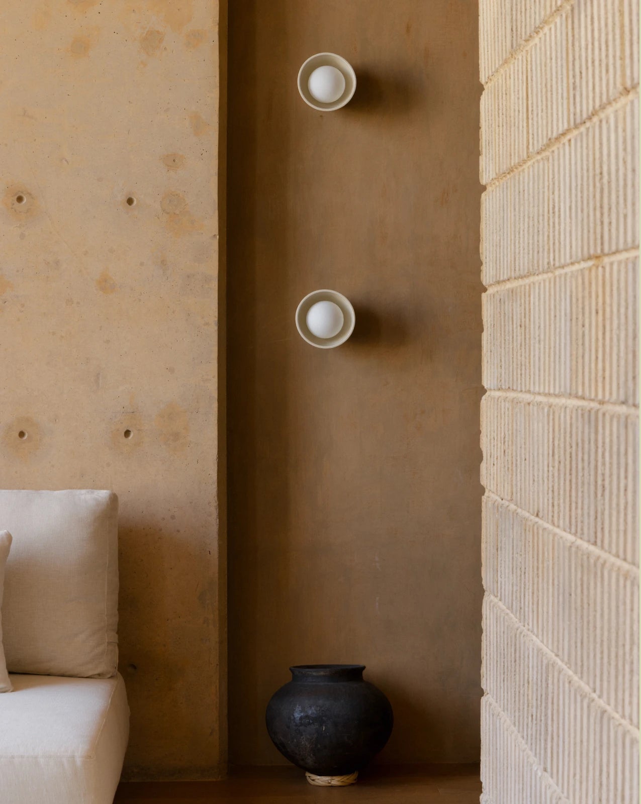 Two modern wall sconces with white circular bulbs mounted vertically on a warm brown plaster wall, flanked by a textured beige brick wall and a cream sofa.