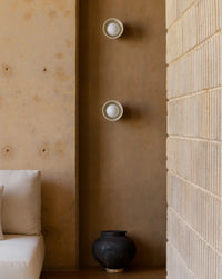 Two modern wall sconces with white circular bulbs mounted vertically on a warm brown plaster wall, flanked by a textured beige brick wall and a cream sofa.