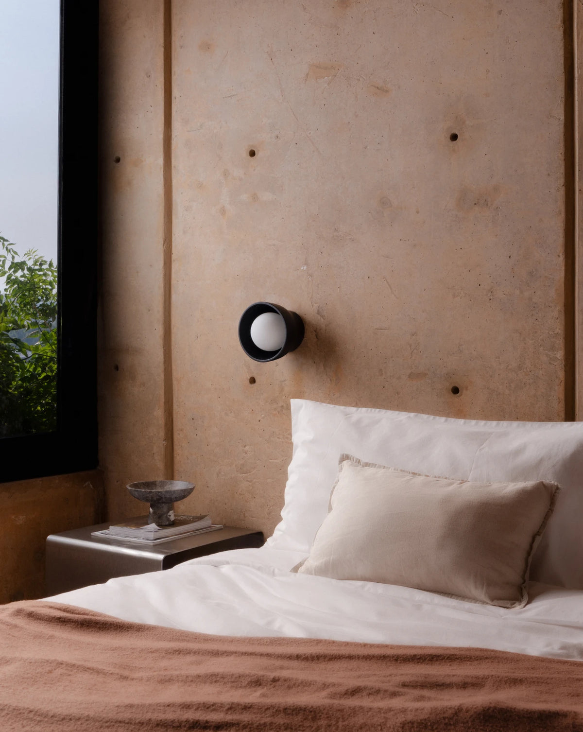 A modern black wall-mounted sconce with a white globe bulb, mounted on a textured concrete wall above a bed with white linens and a brown throw blanket.