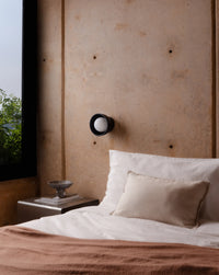 A modern black wall-mounted sconce with a white globe bulb, mounted on a textured concrete wall above a bed with white linens and a brown throw blanket.