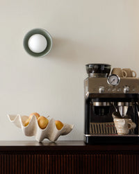 A modern wall-mounted light fixture with a white globe bulb, mounted on a white wall above a dark wood cabinet. On the cabinet sits a ceramic shell-shaped fruit bowl with apples and a countertop espresso machine with ceramic mugs.