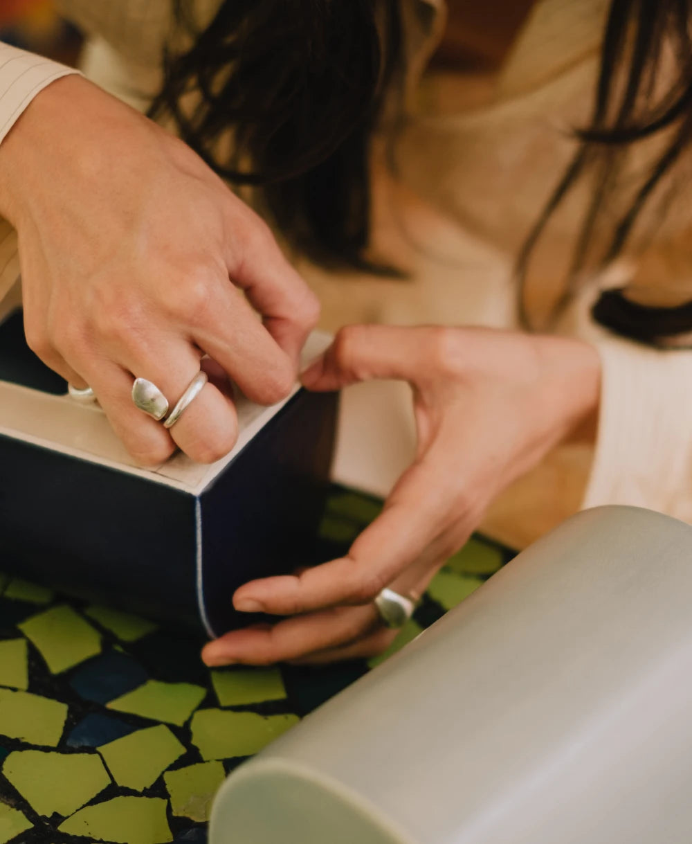 Close-up of a person's hands wearing silver rings while holding a ceramic wall sconce atop a green mosaic surface.